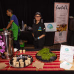 A chef in a black uniform and cap stands behind a table with small cups of soup and fresh greens at a catering event. Signs for "Feed It Forward" and "Capital L" are displayed in the background.