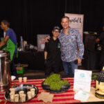 A chef in a black uniform and a man in a colorful shirt pose together behind a catering table with small cups of soup and fresh greens. Signs for "Feed It Forward" and "Capital L" are displayed in the background.