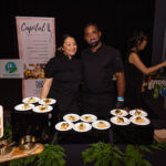 Two chefs in black uniforms stand behind a catering table displaying plated dishes. Signs for "Capital L" and "Feed It Forward" are visible in the background.