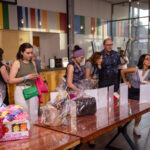 A group of people browses a table filled with wrapped gift baskets at an indoor event. Some individuals are closely examining the items while others converse.