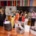 People browse a table with wrapped gift baskets at an indoor event. Some individuals examine the items closely, while others converse or use their phones.