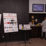A display board thanking food and beverage partners stands next to a small table with promotional items. In the background, a bar counter with a drink station and a staff member is visible.