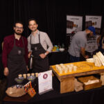 Two men wearing aprons stand behind a food and drink station, smiling at the camera. The table is set with small cups, ingredients, and a display sign. In the background, a man is preparing something at another booth with banners promoting award-winning spirits.
