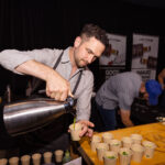 A man wearing an apron pours a green liquid from a metal pitcher into small paper cups on a wooden table. In the background, another person is preparing food at a station with banners promoting award-winning spirits.