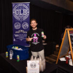 A man in a black Great Lakes Brewery t-shirt smiles and holds up a can of beer while standing in front of a promotional booth with a blue GLB banner. A table with plastic cups, a chalkboard sign, and a bucket filled with canned beverages is set up in front of him.