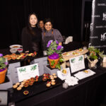 Two women stand behind a table displaying an assortment of small tarts and plated dishes at a food event. The table is decorated with flowers and signs labeling the dishes, including "Summer Tarts" and "Chicken Kofta." A third person is partially visible smiling in the background.