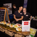 Two women wearing "Hooked" uniforms stand behind a table displaying Vietnamese ahi tuna lettuce wraps. The table is decorated with fresh ingredients and a sign describing the dish. A chalkboard menu and promotional banners are visible in the background.