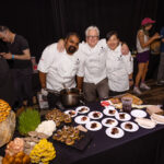 Three chefs in white uniforms stand together behind a table filled with plated gourmet dishes, including appetizers and desserts. Various ingredients, such as mushrooms and microgreens, decorate the table. The chefs smile while posing for the photo.