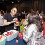 A man in glasses and a black shirt pours a canned beverage into a cup for a woman in a silver outfit at a crowded event. A table with coolers filled with drinks and additional cans is in the foreground.