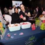 Two people stand behind a table with a "GRUVI" banner, holding canned and bottled beverages. The table has coolers filled with drinks, stacks of cups, and promotional stickers. A crowd mingles in the background at the event.