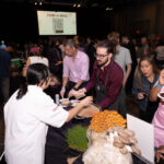 A chef in a white coat serves food to an attendee wearing an apron at a busy event. A table in the foreground displays fresh mushrooms and herbs. A crowd mingles in the background, with a "FUND-A-MEAL" sign projected on the screen.