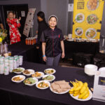 A smiling chef in a black and red uniform stands behind a table with plated dishes, bananas, and food containers at a food event. A colorful sign in the background showcases various meal options.