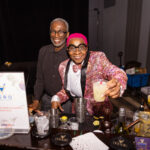 Two bartenders stand behind a bar at an event, one wearing a black outfit and the other in a vibrant pink suit with matching hair, holding up a cocktail with a big smile. Various drink-making ingredients and tools are on the counter.