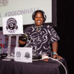 A DJ wearing headphones and a black patterned shirt smiles behind a DJ booth with a laptop and mixer at an event. A sign with social media handles is displayed on the table.