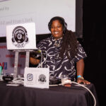 A DJ wearing headphones and a black patterned shirt smiles while performing at an event. A laptop with a "Dreadie Love" logo is on the DJ booth, along with a mixer and a sign displaying social media handles.
