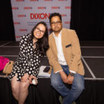 A woman in a polka dot dress and a man in a tan blazer sit on the edge of a stage, smiling at the camera. A "DIXON" banner is visible in the background.