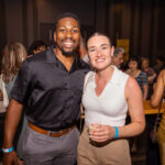 A smiling man in a black shirt and a woman in a white top pose together at a crowded event, holding drinks.