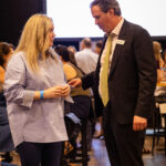 A man in a suit and a woman in a blue blouse are engaged in conversation at a crowded event.