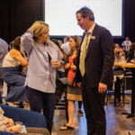 A man in a suit and a woman in a blue blouse are engaged in conversation at a busy event, with people socializing in the background.