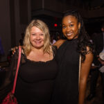 Two smiling women in black outfits pose together at an event, with people mingling in the background.