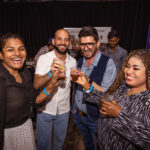 A group of four people is smiling and raising their drinks for a toast at an event. They are dressed in casual and semi-formal attire, wearing wristbands. The background features other attendees, booths, and banners, indicating a lively gathering.