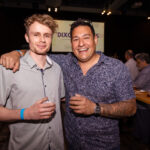 Two men smiling and posing together at an event, each holding a drink, with a "DIXON" sign visible in the background.