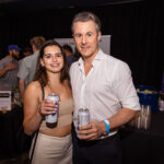 A smiling couple holding canned drinks at an event, surrounded by other attendees.