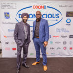 Two men in suits posing in front of a "DIXONLICIOUS" event backdrop featuring various sponsor logos.