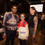 Three people posing at an event, one holding a Dixon Hall tote bag, with a busy background of attendees and a screen displaying auction donors.