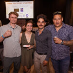 Four people posing at an event, holding drinks and a small plate of food, with a screen in the background displaying event sponsors.
