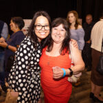 Two women smiling and posing together at an event, one wearing a black and white polka dot dress and the other in a red dress, holding a drink.