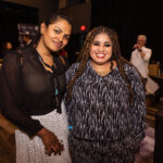 Two women smiling and posing together at an event, one wearing a black and white blouse with matching pants and the other in a black sheer top with a white skirt.
