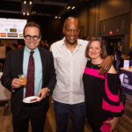 Three people posing together at an event, dressed in semi-formal attire, smiling at the camera while holding drinks and a small plate of food.