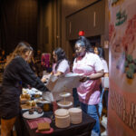 A bakery booth at an event, with staff in pink aprons packing cupcakes into boxes for a customer. The table is decorated with desserts, plates, and promotional materials.