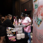 A bakery booth at an event, where staff in pink aprons are packaging cupcakes for a customer. The table is filled with desserts, plates, and a promotional banner displaying colorful macarons and pastries.