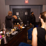 A bartender in a black shirt performs a cocktail smoking technique using a handheld device, creating a smoke-filled bubble over a drink, while guests watch at an event.