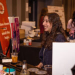 A smiling woman with long curly hair stands behind a bar counter at an event, engaging with guests.