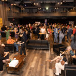 A large event space filled with people socializing, eating, and enjoying drinks, with lounge seating and high tables scattered throughout.