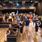 A large event space filled with people socializing, standing around high tables, and sitting on black lounge chairs, with a bustling atmosphere.