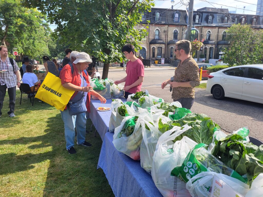 People gathering at an outdoor farmers market, with a woman carrying a yellow shopping bag selecting fresh produce from a table filled with bags of leafy greens. Others are seen in the background engaging in conversation.