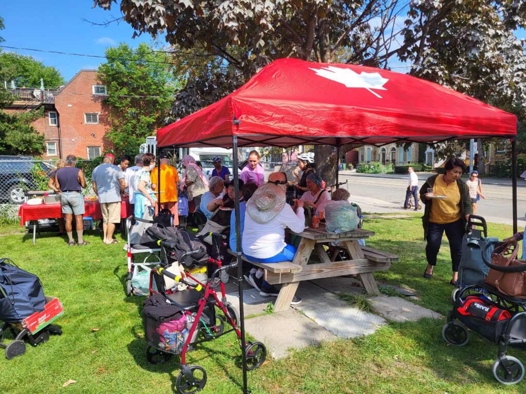 Community members gather outdoors under a red canopy with a Canadian flag, sitting at picnic tables and engaging in conversation. Others stand in line at a food table with red tablecloths, while some walk around with plates of food. Several mobility aids and carts are visible, indicating accessibility considerations.