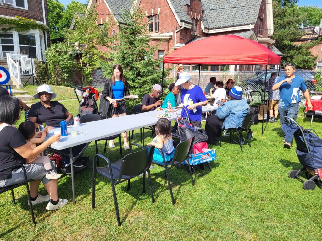 People of various ages and backgrounds gather outdoors at a community event, sitting at tables, eating, and socializing under a red canopy. A woman in a black outfit walks by carrying a plate of food, while others engage in conversation in the grassy area.