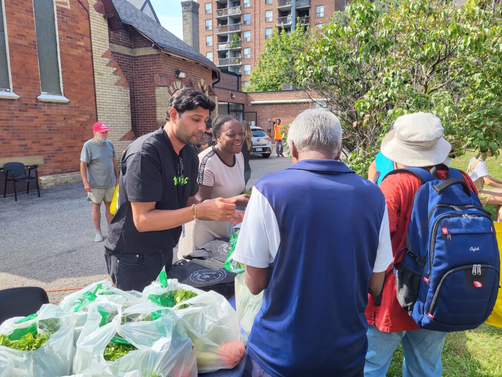 Two volunteers distribute fresh produce in plastic bags to community members at an outdoor event near a brick building, with people waiting in line and engaging in conversation.