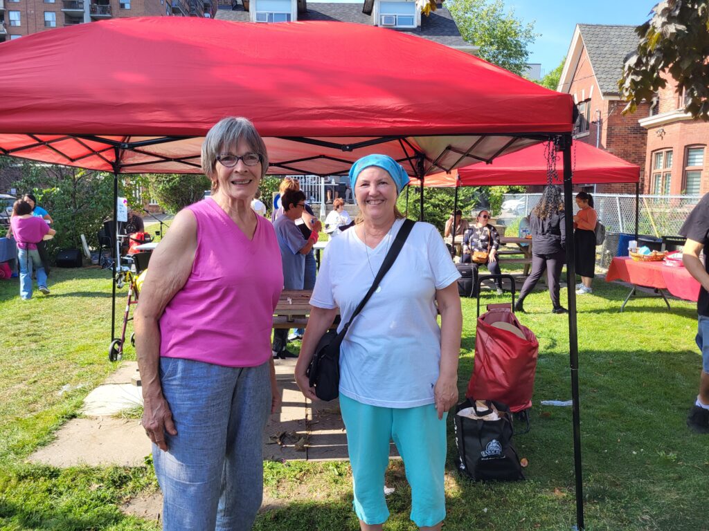 Two smiling women stand in front of a red canopy tent at an outdoor community event, with people gathered in the background sitting at picnic tables and engaging in activities.