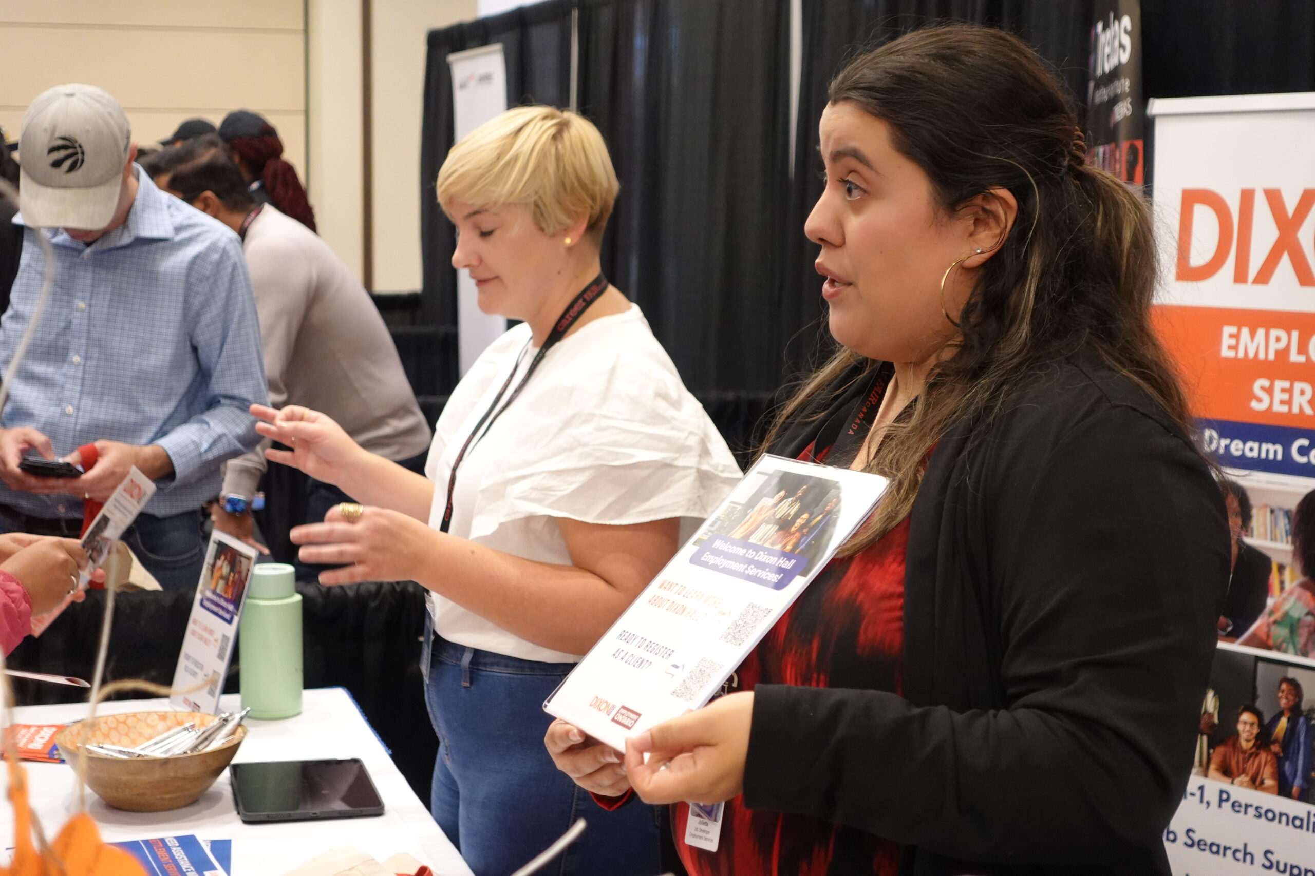 Two women are standing behind a table at a job fair or community event, engaging with attendees. One woman, wearing a black and red outfit, is holding a flyer and speaking to someone. The other woman, wearing a white blouse, is also interacting with visitors. The table is covered with informational materials, a bowl, and a tablet. In the background, a banner with the text "DIXON" and "EMPLOYMENT SERVICES" is visible, along with other attendees and booths.