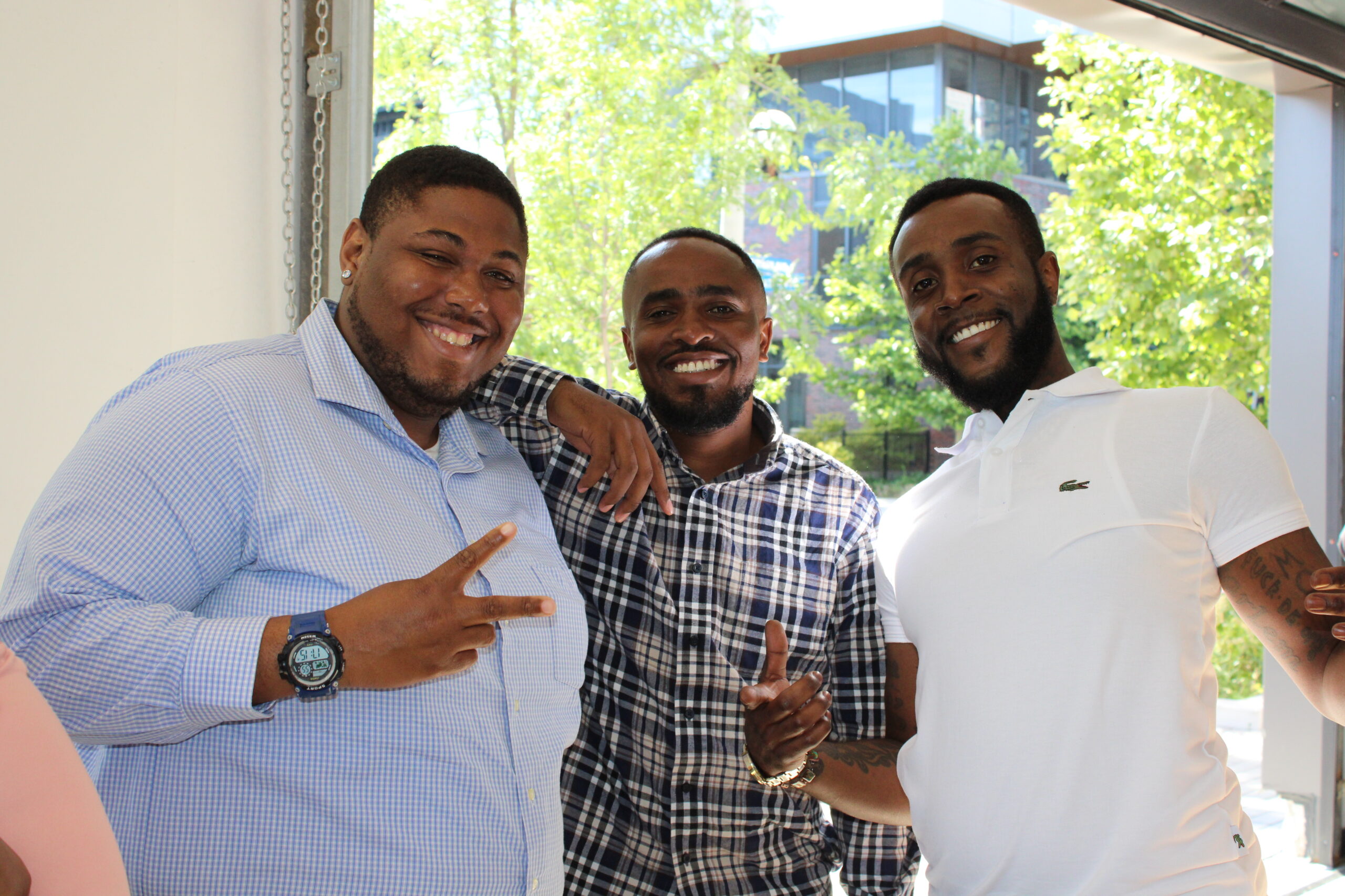 Three men posing together indoors with big smiles. The man on the left is wearing a light blue checkered shirt and making a peace sign, the man in the middle is in a plaid shirt with his arm around the others, and the man on the right is in a white polo shirt, also smiling. They are standing in front of a bright window with trees and a modern building visible in the background.