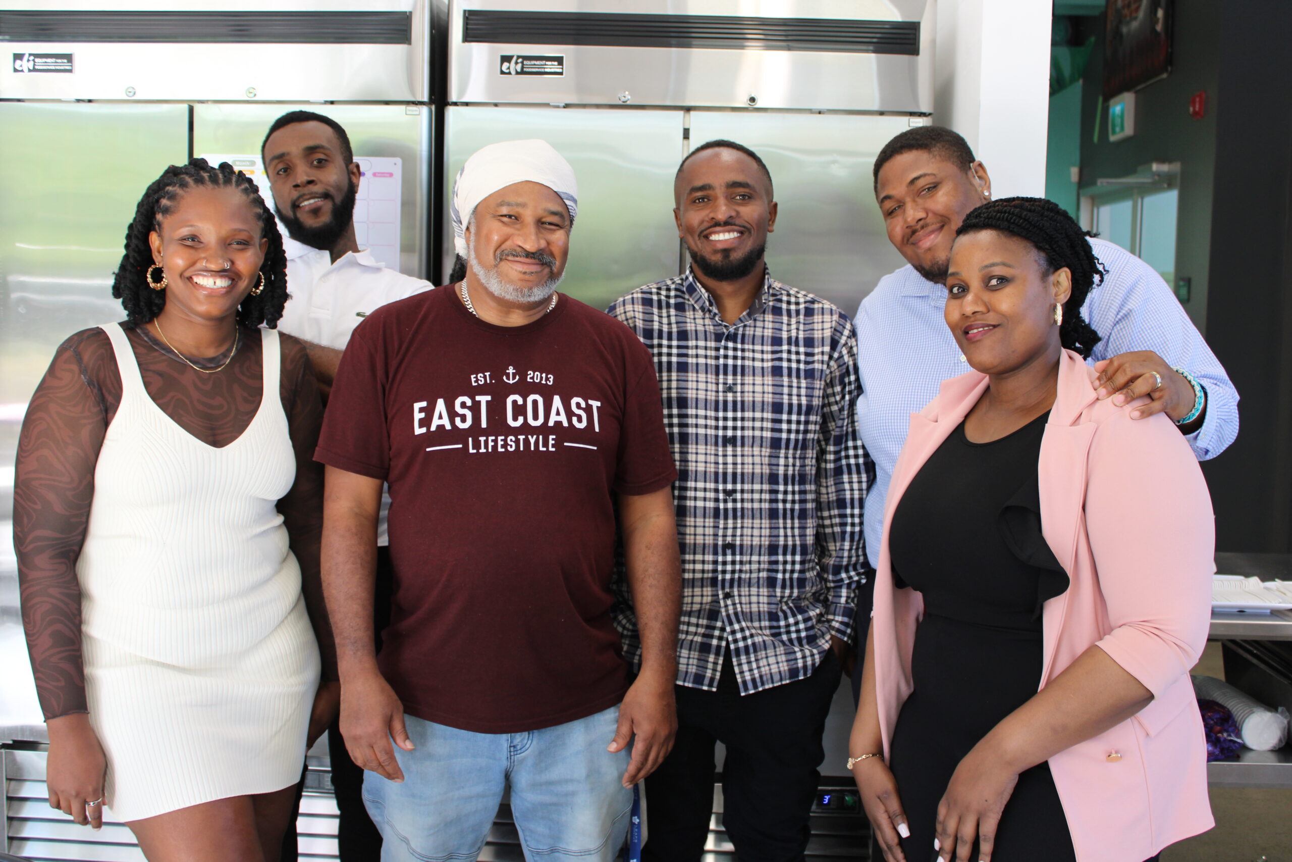 A group of six people is posing together in a commercial kitchen, smiling at the camera. They are diverse in age and style, with some dressed casually and others in business attire. The background features stainless steel refrigerators and kitchen equipment. The group appears to be in a friendly and professional setting.
