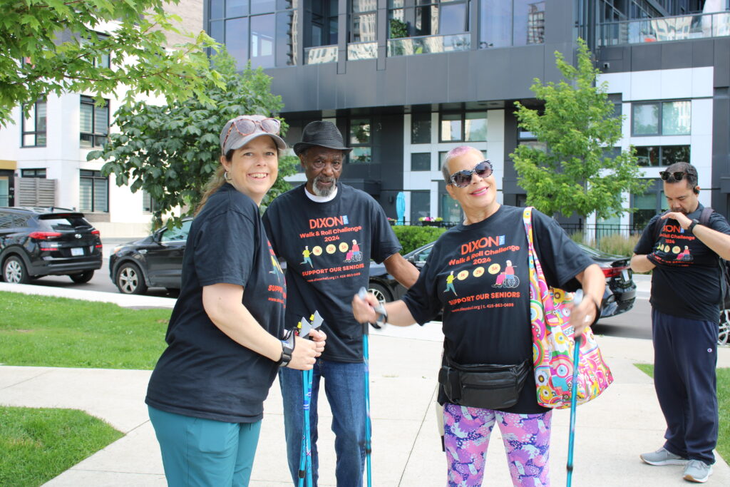 a-group-of-people-wearing-matching-black-t-shirts-with-dixon-walk-roll-challenge-2024-printed-on-them-stand-outdoors-in-an-urban-park-they-are-holding-walking-poles-and-smiling-a-modern-building-with-large-windows-is-visible-in-the-background-