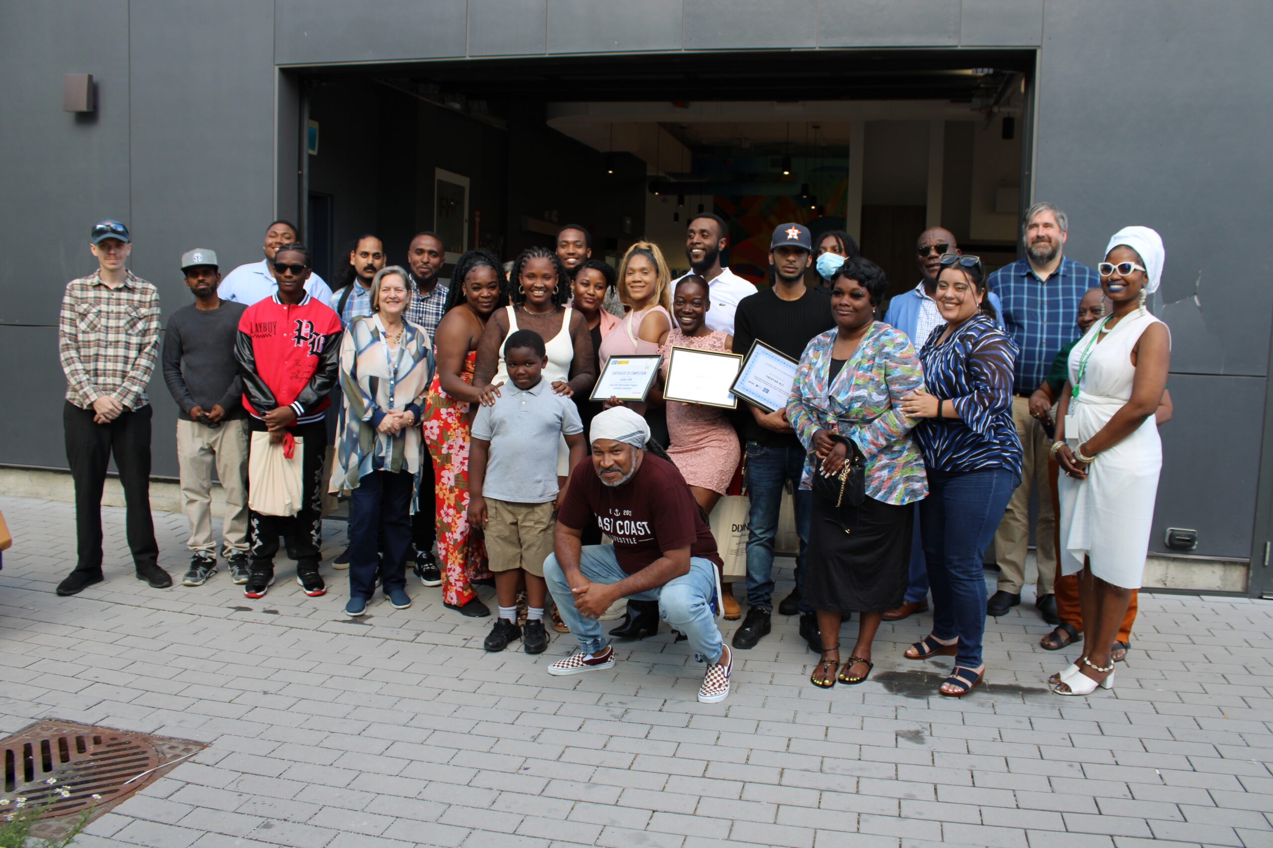 A large group of diverse people gathered outside a building, posing for a group photo. Some individuals in the front row are holding certificates, while others are smiling and embracing each other. A man in a maroon shirt and headscarf is crouching in the foreground. The background features a modern building entrance with colorful artwork inside.