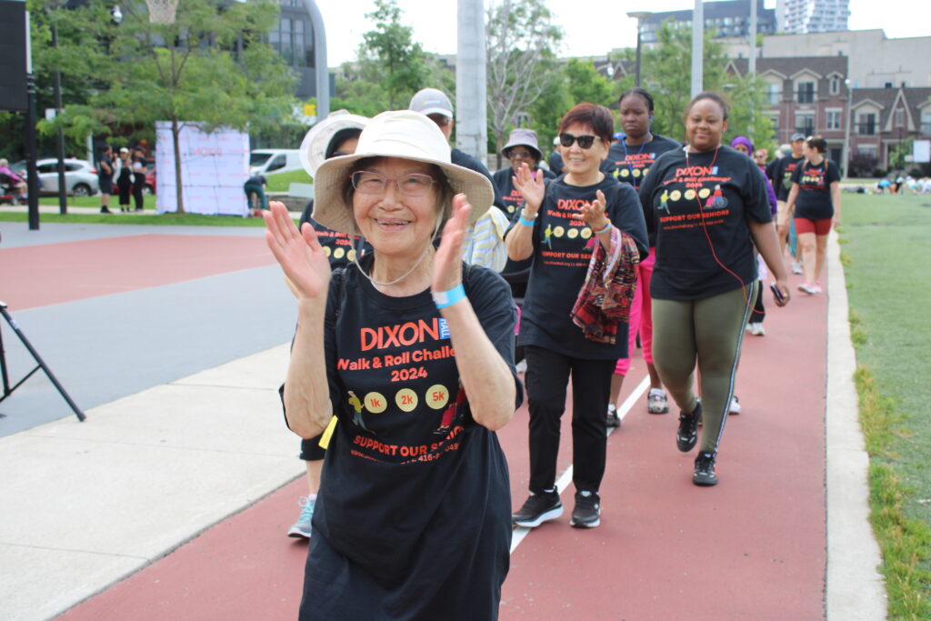a-group-of-people-is-participating-in-the-dixon-walk-roll-challenge-2024-walking-along-a-track-the-leading-woman-wearing-a-hat-and-glasses-is-clapping-and-smiling-other-participants-in-matching-event-t-shirts-follow-behind-some-wearing-headphones-and-sunglasses-the-background-features-a-park-a-basketball-court-and-urban-buildings-